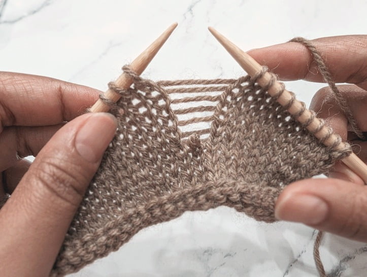 two hands holding knitting needles with a dropped stitch between them, located several stitches down